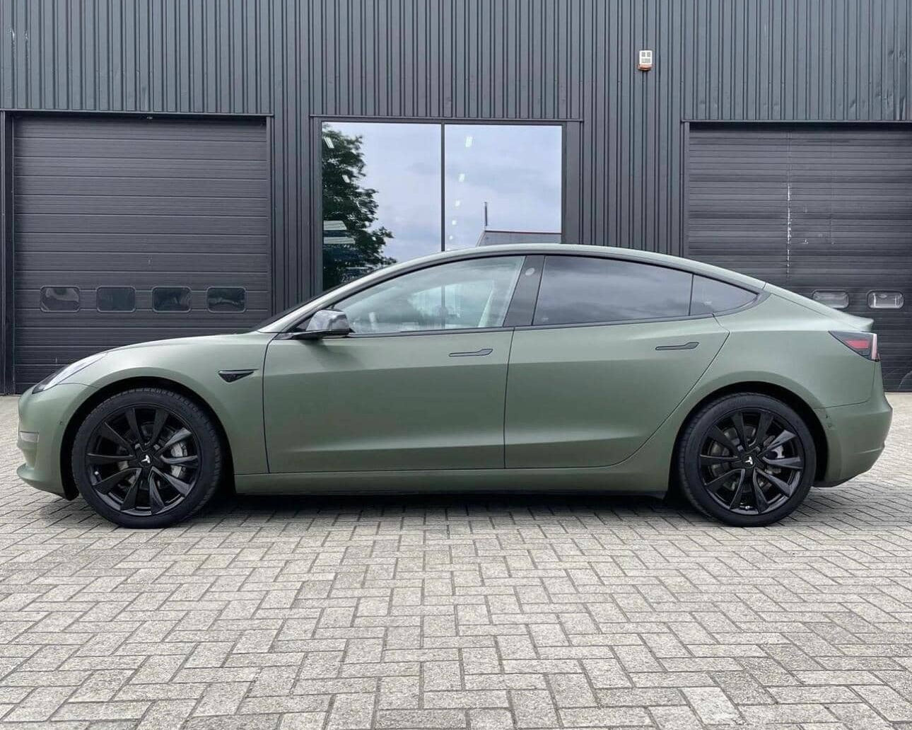 Matte green Tesla Model 3 with black wheels parked on brick pavement outside a modern black building