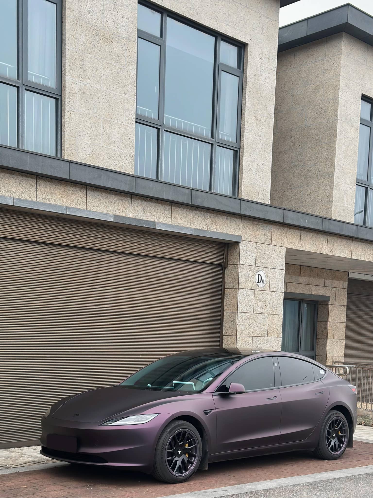 WrapBug matte purple Tesla Model 3 parked in front of a modern garage with black rims