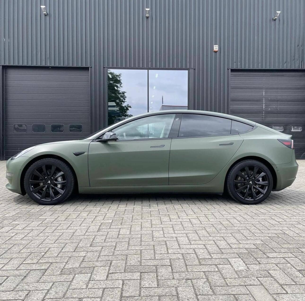 Matte green Tesla Model 3 with black wheels parked on brick pavement outside a modern black building