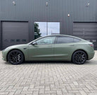 Matte green Tesla Model 3 with black wheels parked on brick pavement outside a modern black building