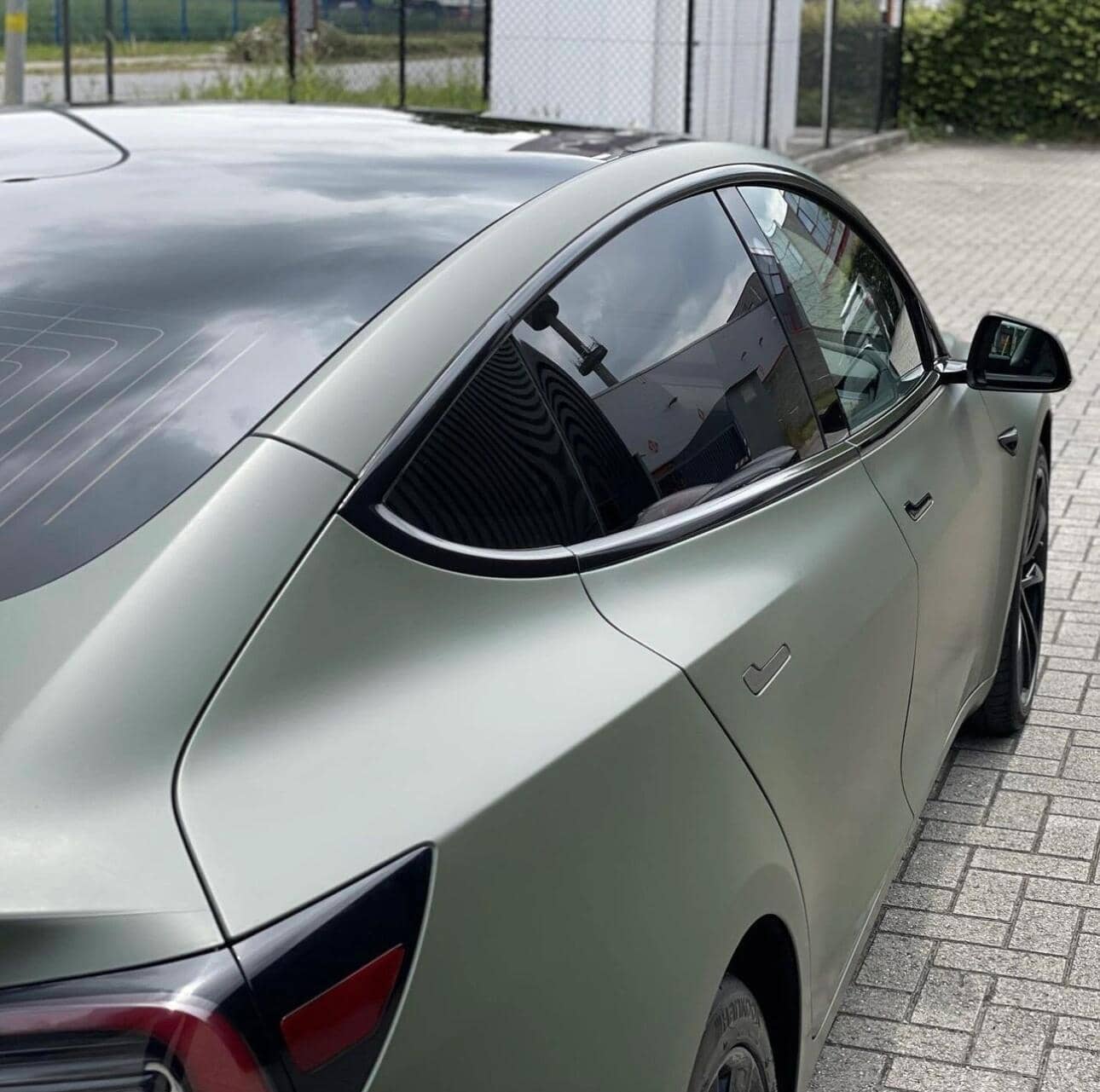 Gray Tesla Model 3 electric car parked on driveway showing rear side and tinted windows