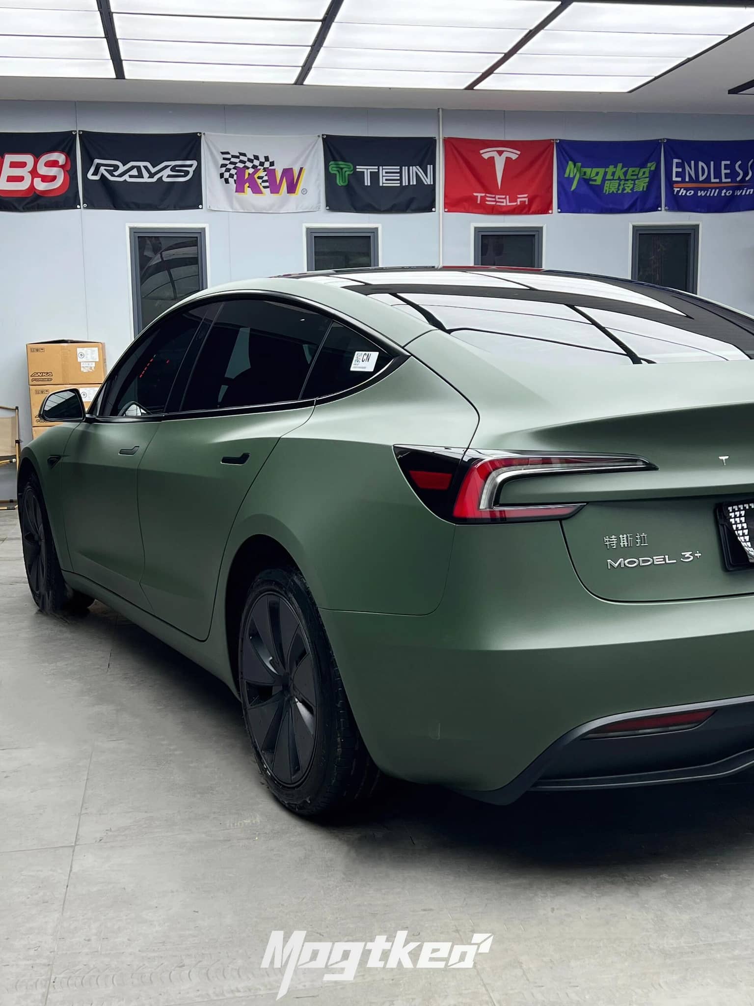 Tesla Model 3 with matte OLIVE GREEN tesla wrap by WrapBug parked indoors with branded banners